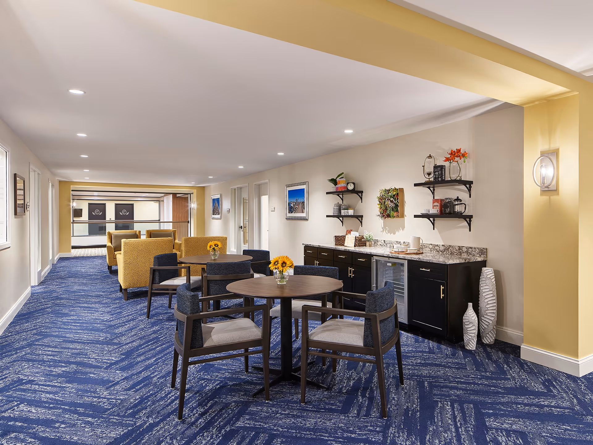 A bright and spacious senior living common area with blue patterned carpet, yellow walls, and recessed ceiling lights. The room features round tables with chairs, small vases of sunflowers on the tables, and a seating area with yellow armchairs. Along one wall, there is a countertop with cabinets, shelves holding decorative items, and a small refrigerator. The space is clean, modern, and inviting.