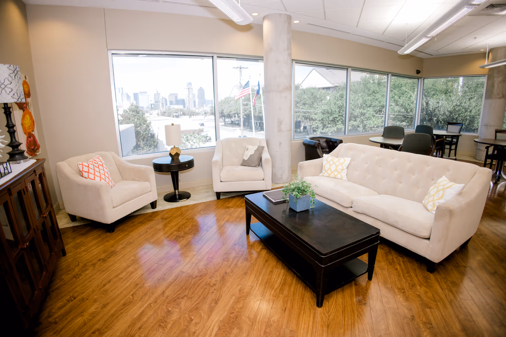 A bright and spacious living room area with large windows showing a cityscape and trees outside. The room features a beige sofa with yellow and white pillows, two beige armchairs with patterned pillows, a dark wooden coffee table with a small plant and book on it, a round side table with a lamp, and wooden flooring. There are additional tables and chairs in the background near the windows.