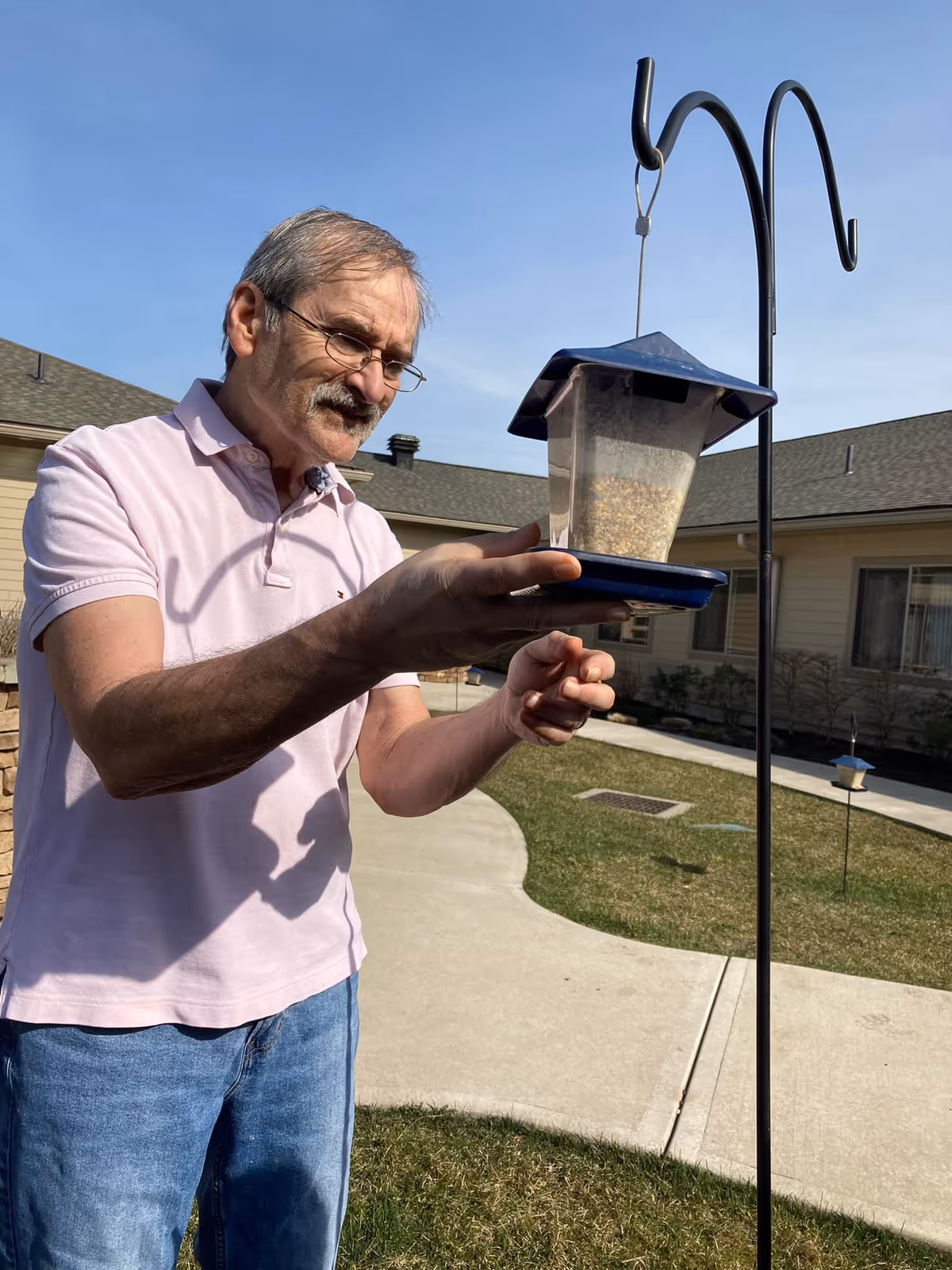 An elderly man wearing glasses, a light pink polo shirt, and blue jeans is outdoors adjusting a blue bird feeder hanging from a black metal stand. The background shows a courtyard with grass, a concrete walkway, and a beige building under a clear blue sky.