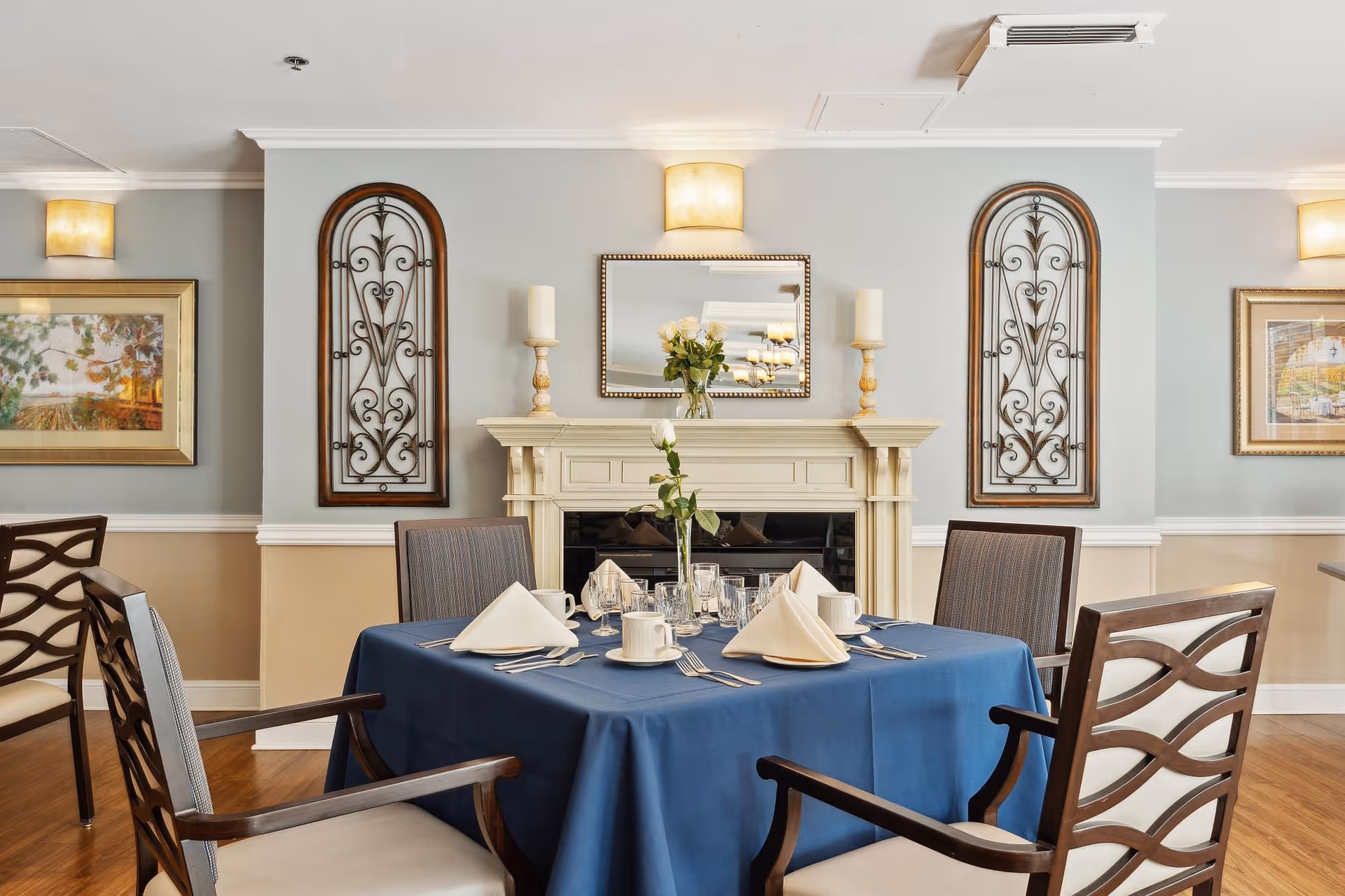 A dining room table set for four with a blue tablecloth, white napkins folded in a triangle, cups, glasses, and silverware. The room features a decorative fireplace with candles and a vase of flowers on the mantel, two ornate wall decorations on either side, framed artwork, and soft wall lighting.