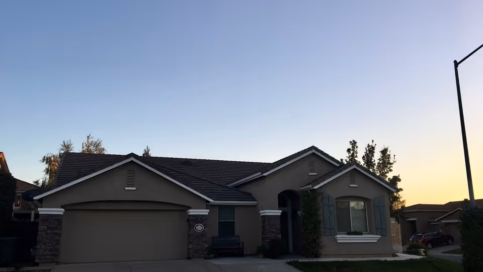 Exterior view of a single-story residential building with a two-car garage, stone accents on the facade, a small front porch with a bench, and a window with decorative shutters. The sky is clear with the sun setting in the background.