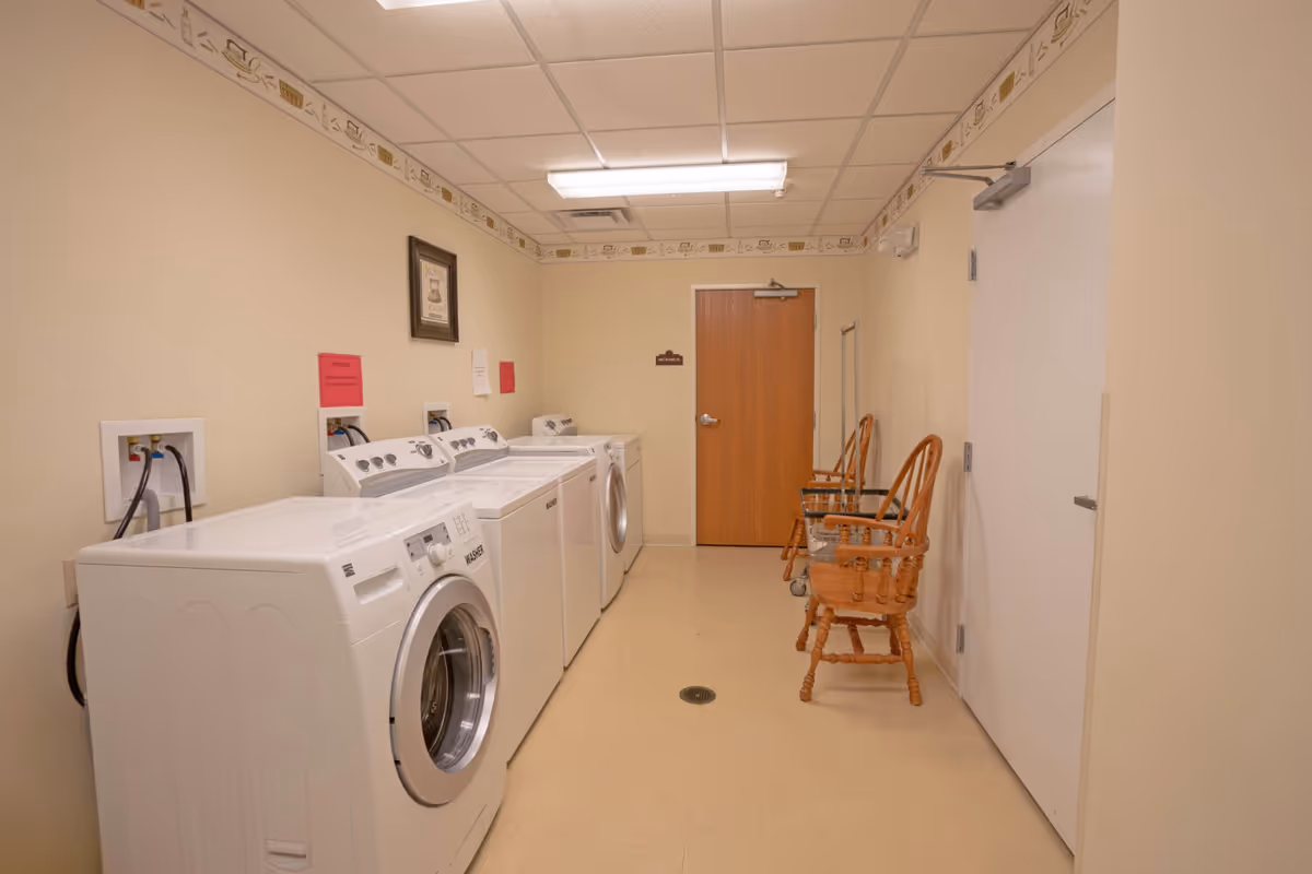 Laundry room with four white washing machines and dryers lined up against a beige wall. Two wooden chairs and a laundry cart are placed along the opposite wall. The room has a beige floor, a drop ceiling with fluorescent lighting, and a wooden door at the far end.