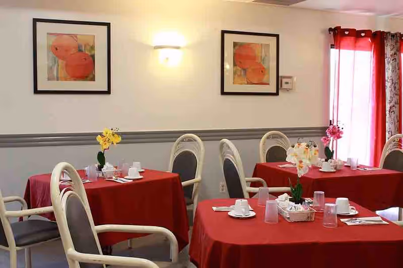 Small dining room with red tablecloth-covered tables, chairs, floral centerpieces, and framed wall art on the wall.