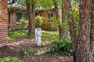 Small garden courtyard with trees, plants, a brick building, and a white angel statue.