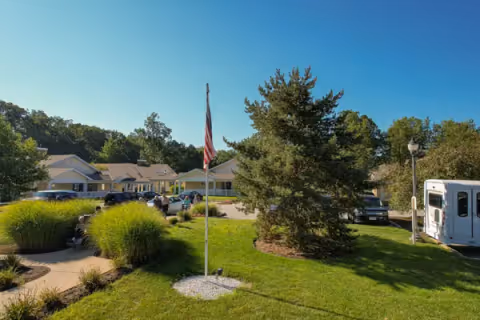 Exterior view of Maple Ridge Senior Living facility showing a well-maintained lawn with a flagpole displaying the American flag, surrounded by trees and bushes. Several cars are parked near the single-story building under a clear blue sky.