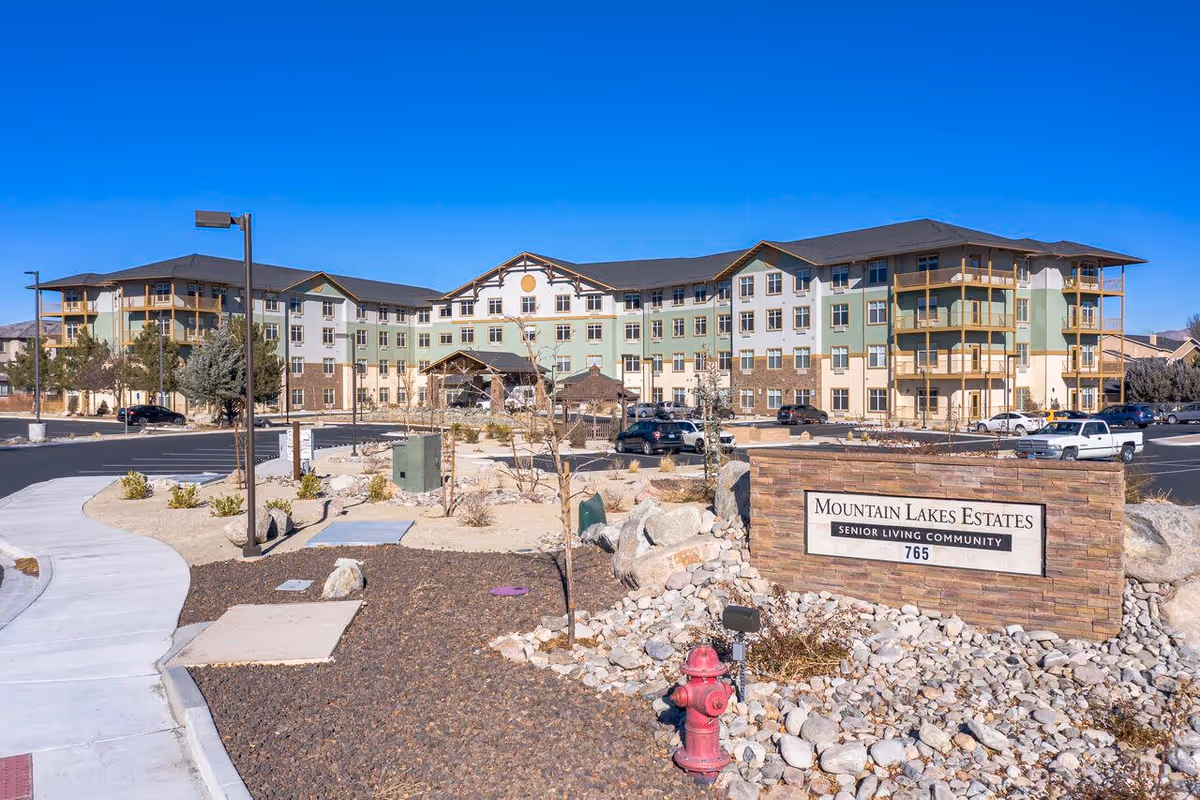 Exterior view of Mountain Lakes Estates, a senior living community building with multiple floors, balconies, and a parking lot in front. The foreground shows a landscaped area with rocks, a fire hydrant, and a stone sign displaying the facility name and address.