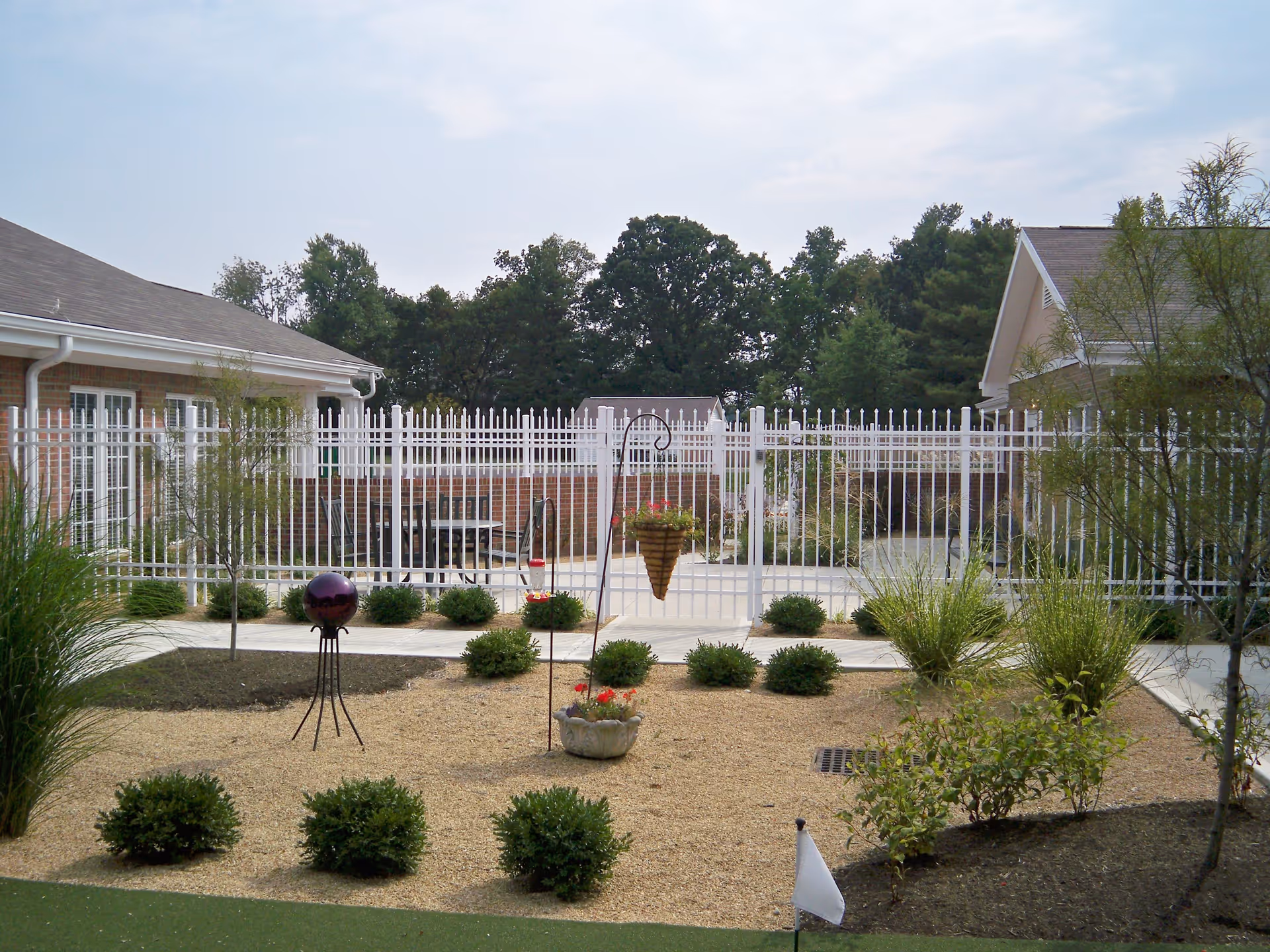Outdoor garden area at Morrison Woods Health Campus featuring a gravel bed with small bushes and ornamental plants, a decorative purple sphere on a stand, a hanging flower basket, and a hummingbird feeder. The garden is enclosed by a white metal fence with brick buildings on either side and trees in the background under a partly cloudy sky.