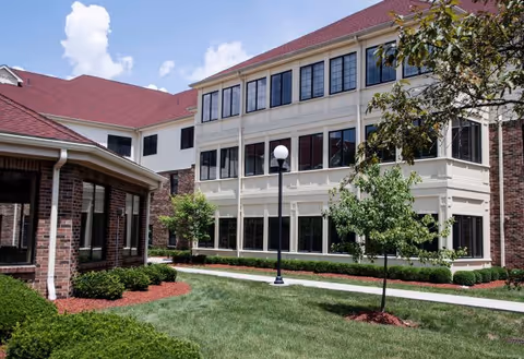 Exterior view of a senior living facility building with red roofs, large windows, and a well-maintained lawn with small trees and shrubs under a partly cloudy sky.