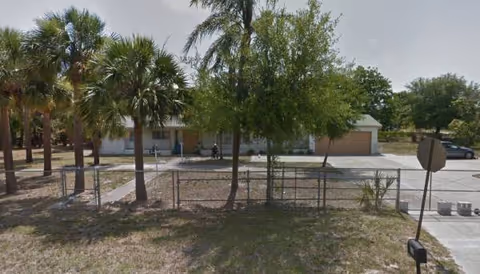 Exterior view of a single-story facility with palm trees, a fence, and a driveway.