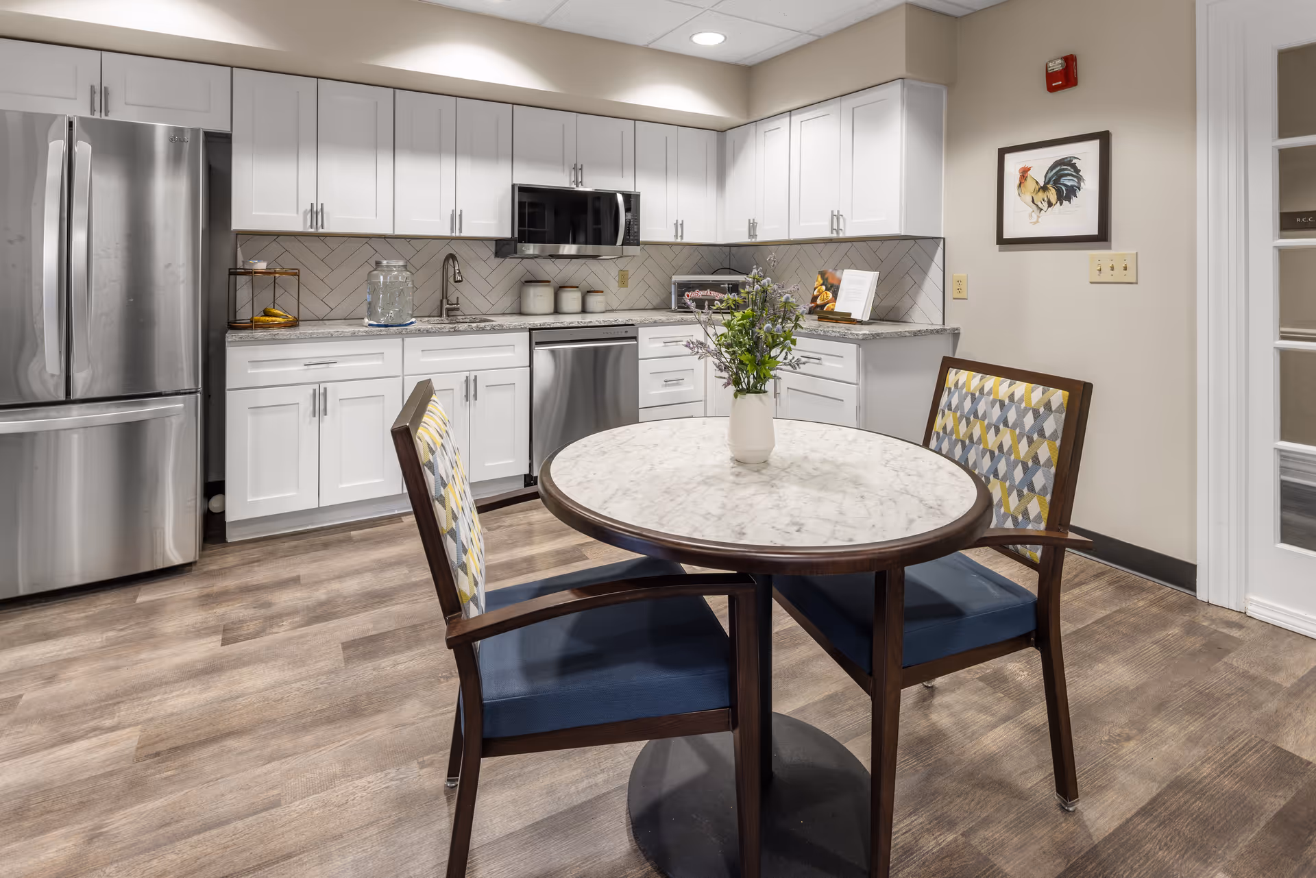 A modern kitchen area with white cabinets, stainless steel refrigerator, dishwasher, and microwave. There is a round table with a marble top and two chairs with patterned backs and blue seats. A vase with flowers is placed on the table. The walls are light-colored with a framed picture of a rooster and a fire alarm mounted above it.