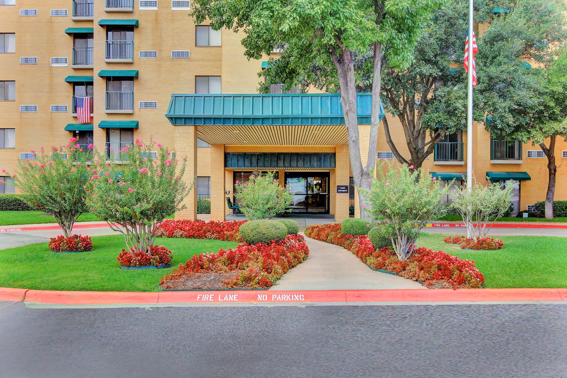 Entrance of a multi-story brick building with green awnings over windows and the main entrance. The entrance has a covered walkway with a green roof and glass doors. There are well-maintained flower beds with red flowers and green bushes lining the walkway, along with several trees. An American flag is visible on a flagpole to the right of the entrance. The curb in front is painted red with white text reading 'FIRE LANE NO PARKING'.