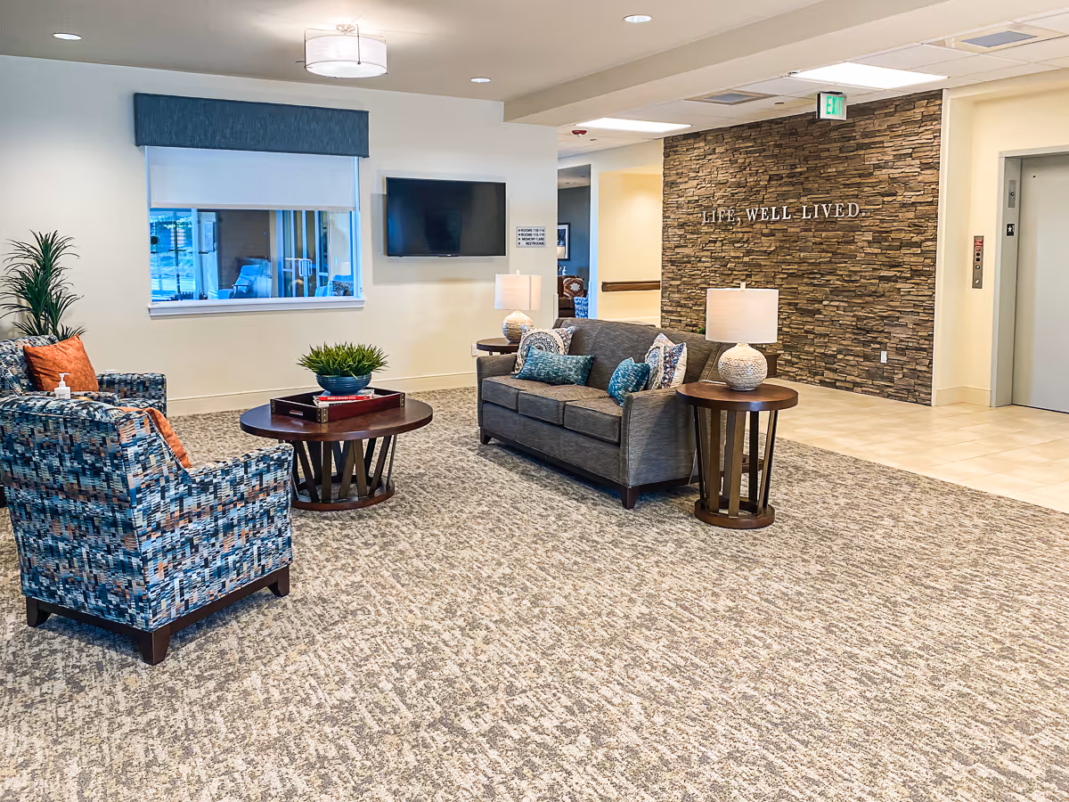 A bright and welcoming assisted living facility common area with a patterned armchair, a gray sofa with decorative pillows, two round wooden side tables with lamps, a round coffee table with a plant and tray, a large window with a blue valance, a wall-mounted TV, and a stone accent wall with the words 'LIFE, WELL LIVED.' An elevator is visible on the right side.