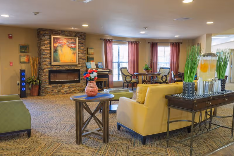 A cozy living room area in a senior living facility featuring a stone fireplace with a colorful painting above it, comfortable seating including a yellow armchair and a green chair, a round wooden table with a vase of flowers, a table with a beverage dispenser filled with lemon water, and large windows with red curtains letting in natural light.