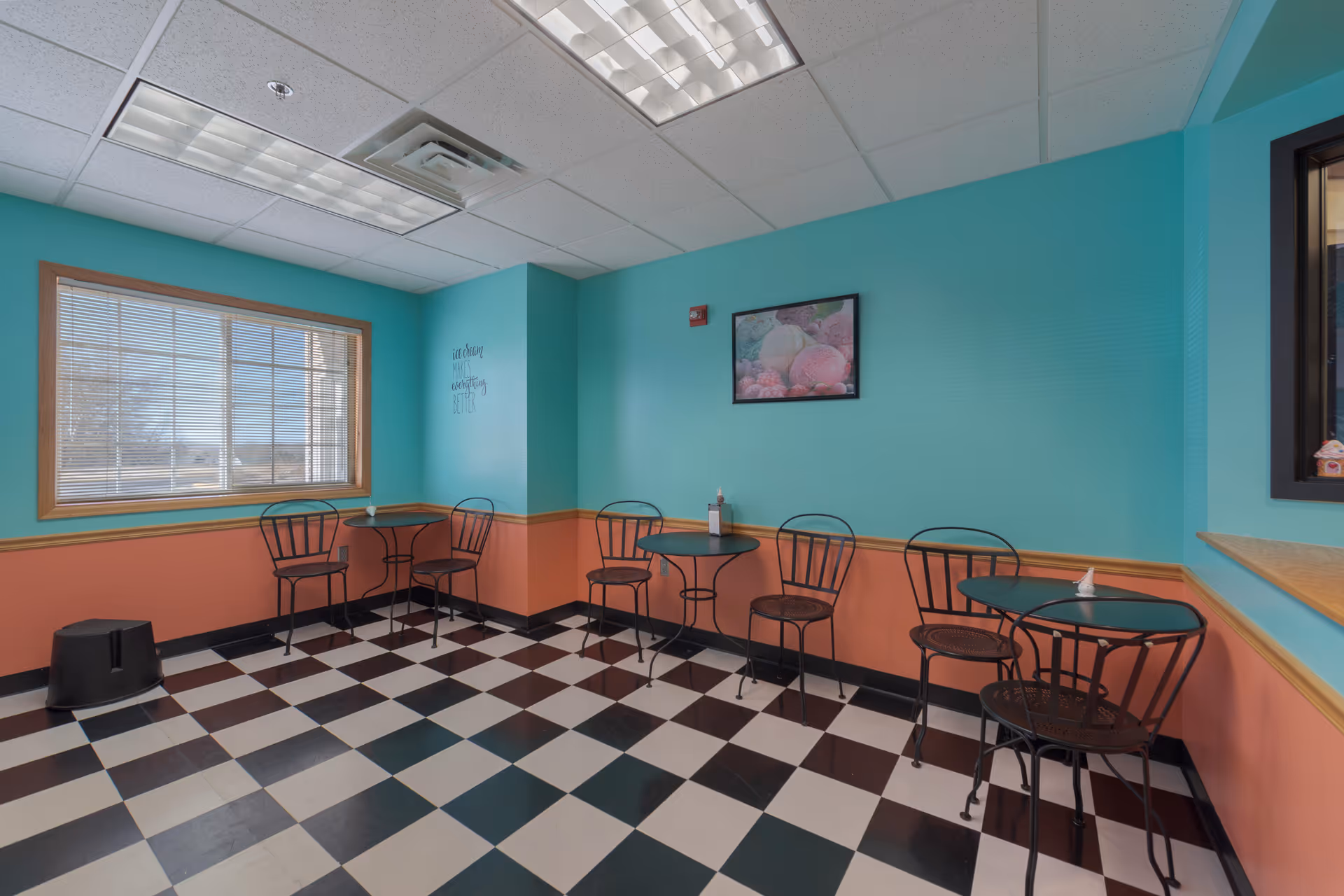 A small dining area with turquoise and peach walls, black and white checkered floor, and three small round tables each with two black metal chairs. A window with blinds lets in natural light, and a framed picture of ice cream cones hangs on the wall.