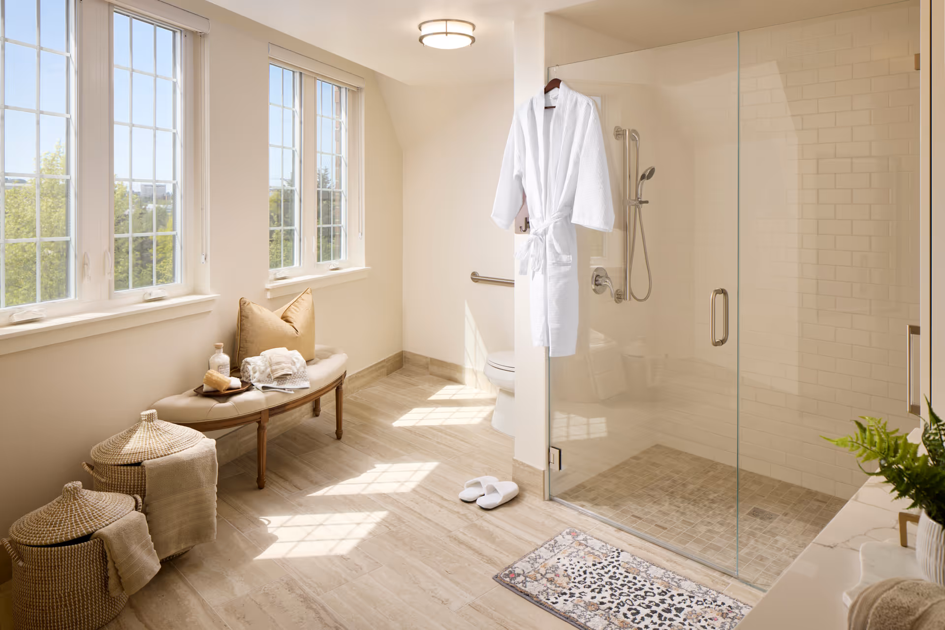 Bright, spacious bathroom with a glass shower, a hanging white robe, a bench by large windows, woven baskets, and slippers on a tiled floor.
