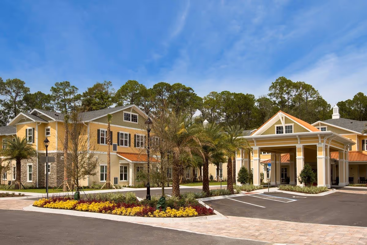 Exterior view of a senior living facility named Starling at Ponte Vedra, featuring a large yellow and beige building with multiple windows, a covered entrance with white columns, palm trees, landscaped flower beds, and a clear blue sky.