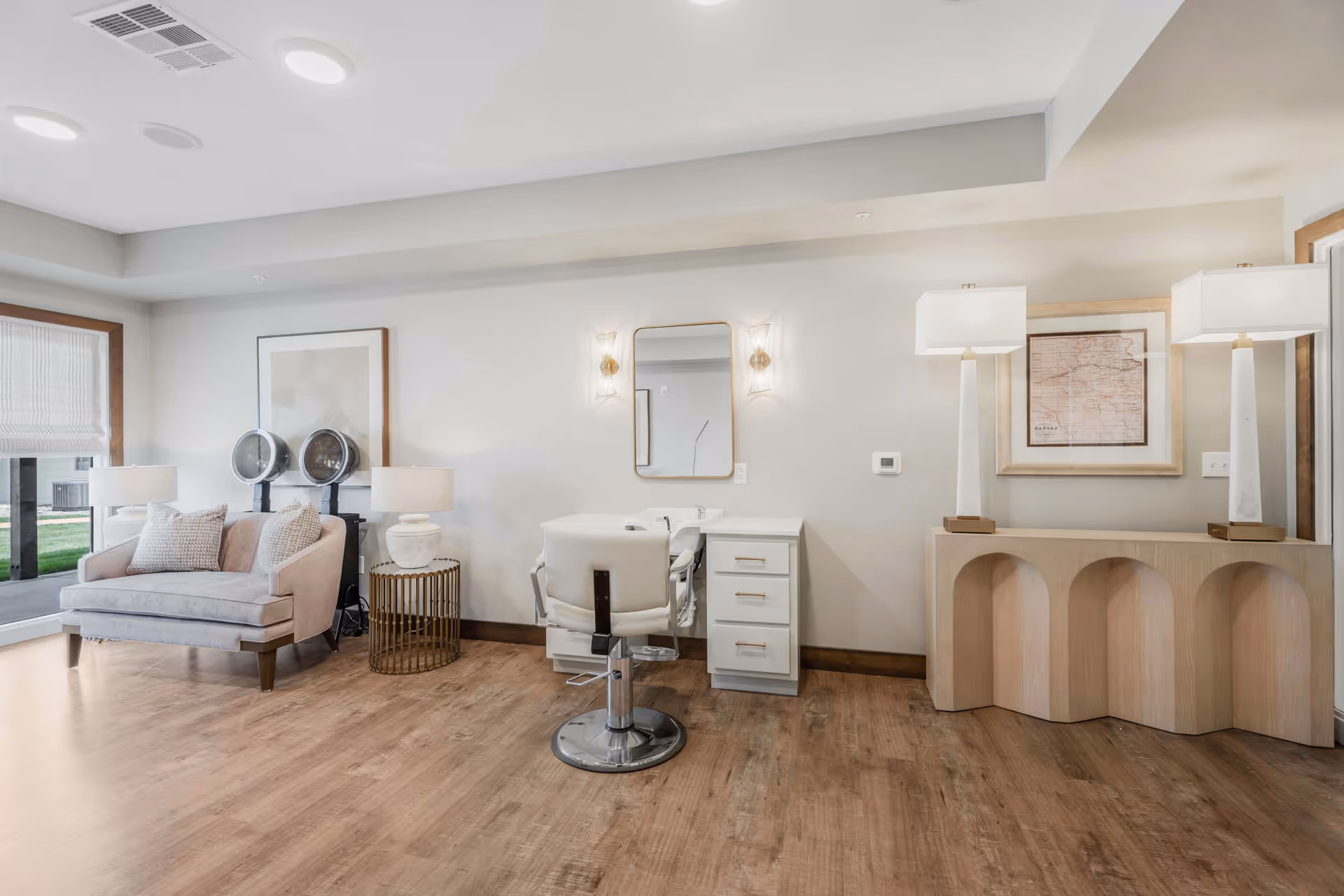 A bright and modern salon area in a senior living facility featuring a styling chair in front of a vanity with a mirror and wall sconces. To the left, there is a comfortable loveseat with two pillows and two hair dryer stations behind it. To the right, a console table with two tall lamps and a framed map on the wall. The room has wood flooring and large windows letting in natural light.