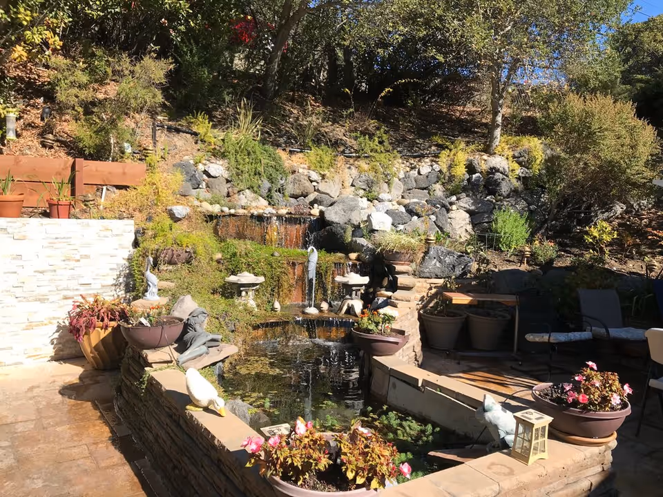 Outdoor garden area with a small pond featuring a multi-tiered waterfall surrounded by rocks and greenery. There are potted plants, garden statues, and seating chairs arranged around the pond. The area is sunlit with trees and shrubs in the background.