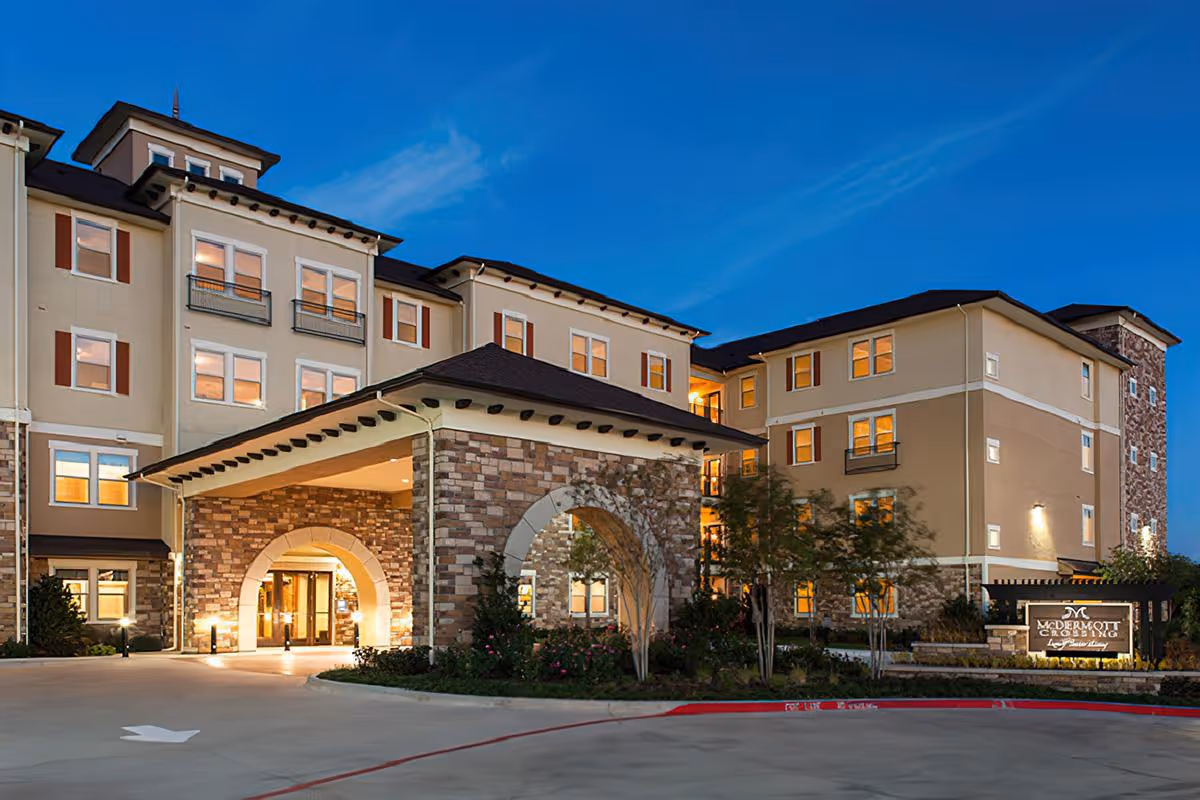 Exterior view of McDermott Crossing senior living facility at dusk, showing a multi-story building with illuminated windows, stone and beige siding, an arched entrance, landscaped greenery, and a sign with the facility name.