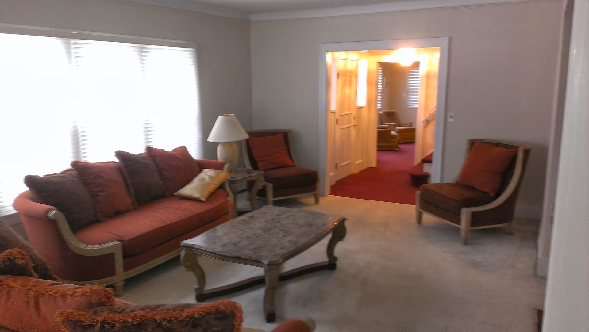 A living room with a red sofa and matching armchairs, a marble-top coffee table in the center, a side table with a lamp, and a large window with blinds letting in bright light. The room has light-colored walls and carpet, and an open doorway leads to another room with red carpet and additional seating.
