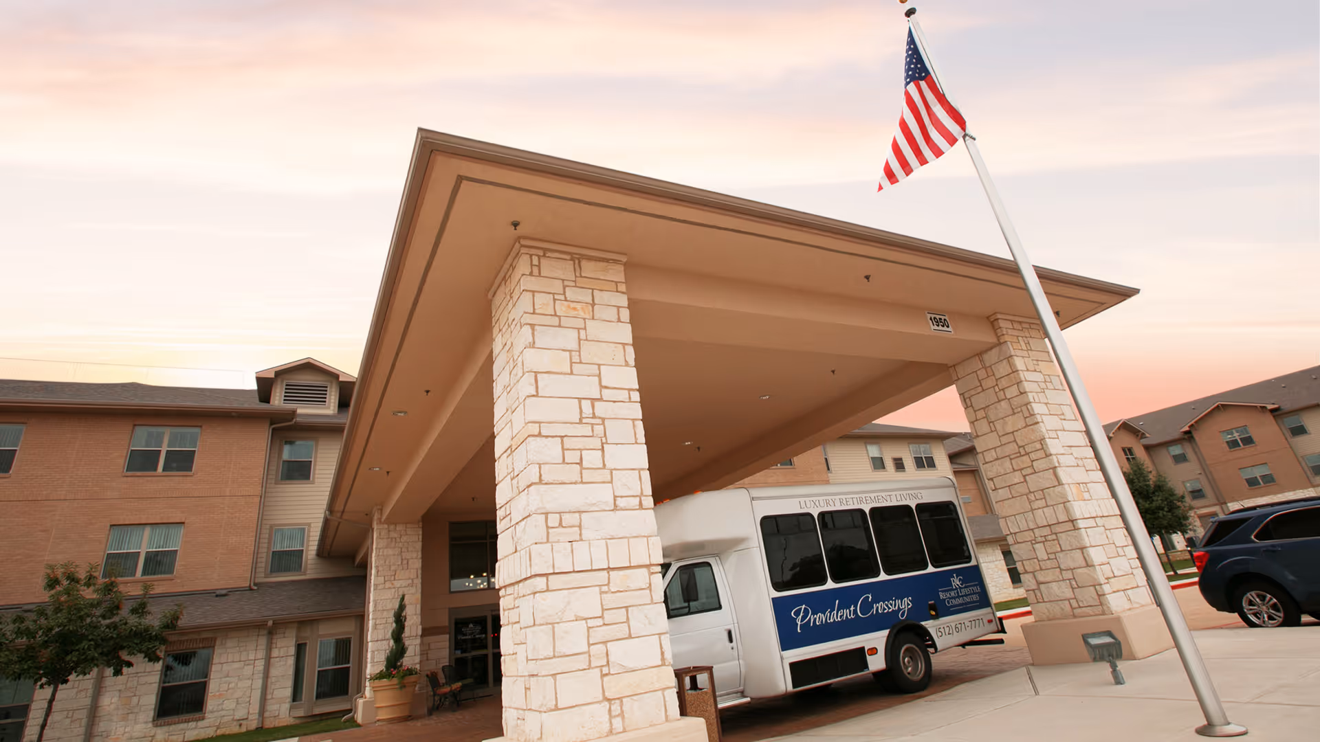 Entrance of Provident Crossings Retirement Resort showing a covered drop-off area with stone pillars, an American flag on a pole, and a white shuttle bus parked under the canopy. The building is a multi-story structure with beige and stone exterior walls.