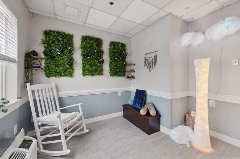 A cozy corner of a room with a white rocking chair, three vertical green plant wall decorations, a dark wooden bench with cushions, a tall white floor lamp, and decorative hanging plants on a light gray and blue wall.