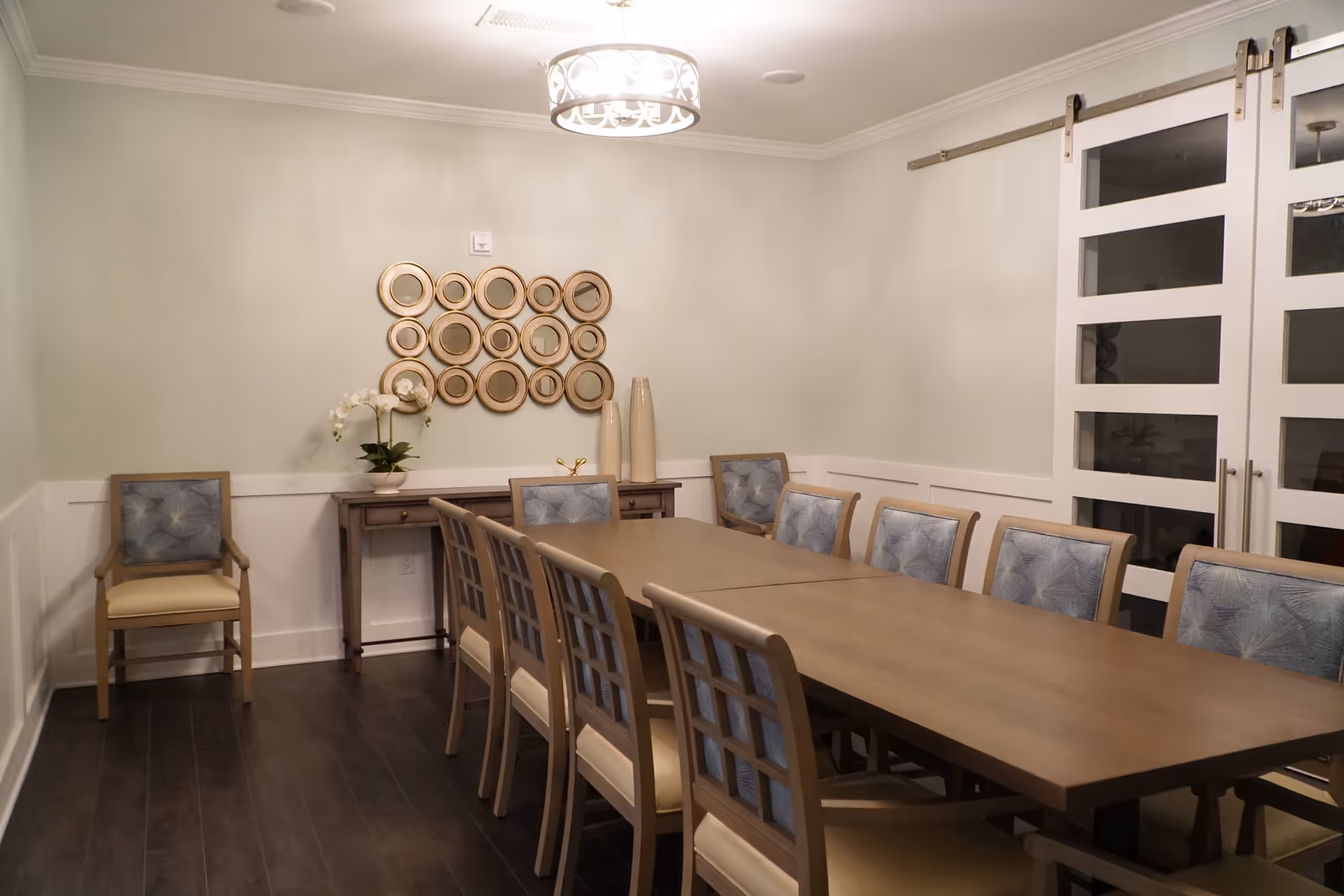 A dining room with a long wooden table surrounded by ten chairs with light blue patterned cushions. There is a single chair with a similar cushion against the left wall. On the back wall, there is a wooden console table with decorative vases and a potted orchid. Above the console table, there is a wall decoration made of multiple round mirrors with gold frames. The room has light-colored walls, dark wooden flooring, a modern ceiling light fixture, and white sliding barn doors with glass panels on the right side.