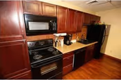 Interior view of a kitchen featuring dark wood cabinets, a black microwave mounted above a black electric stove, a coffee maker on the countertop, a dishwasher, and a refrigerator. The floor is wooden and the walls are painted a light color.