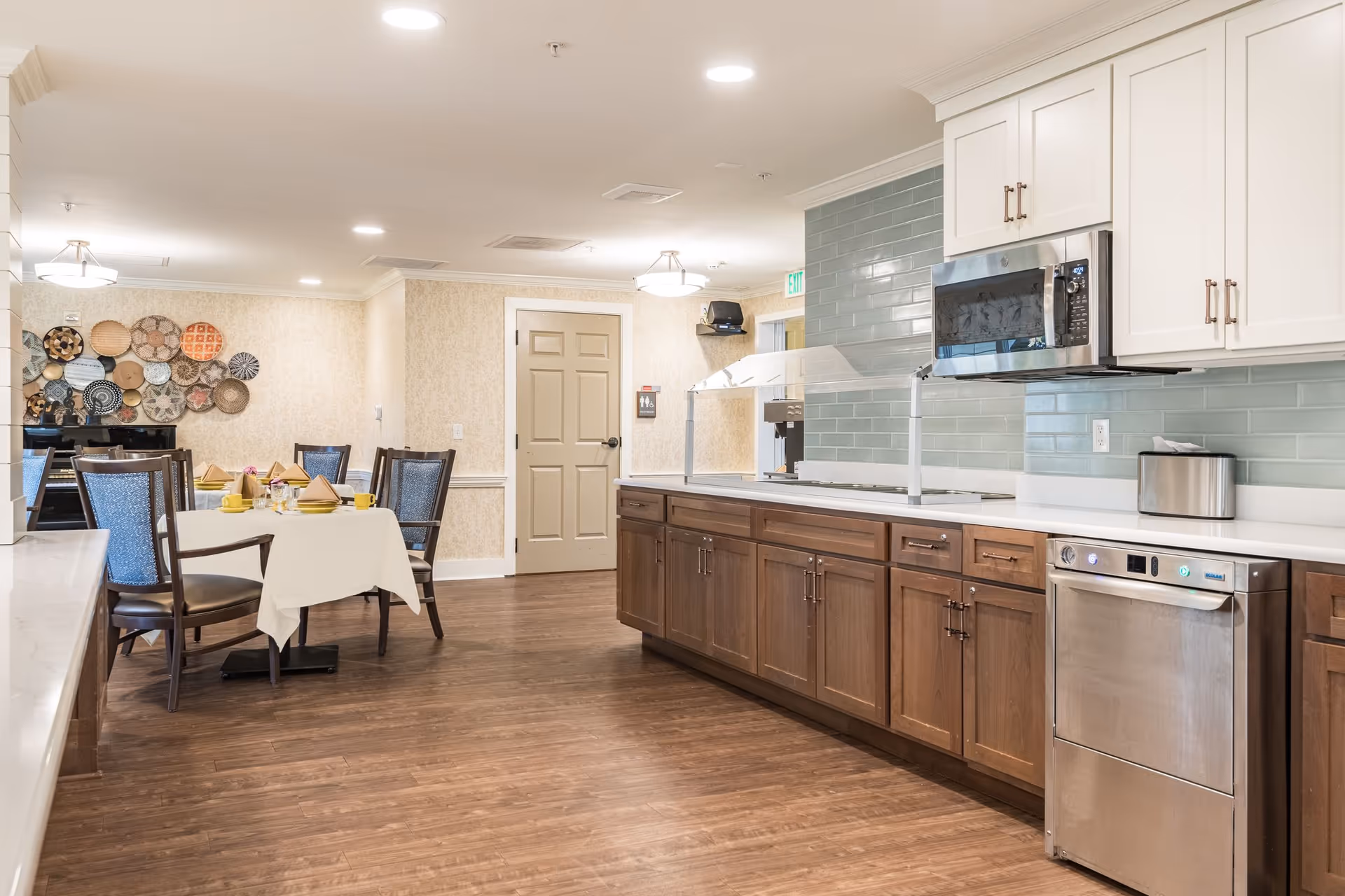 A clean and well-lit dining area and kitchen in a senior living facility. The kitchen features wooden cabinets, a stainless steel microwave, and dishwasher, with a light blue tiled backsplash. The dining area has a table set with yellow cups, plates, and folded napkins, surrounded by wooden chairs with blue cushions. Decorative woven baskets are mounted on the wall in the background.