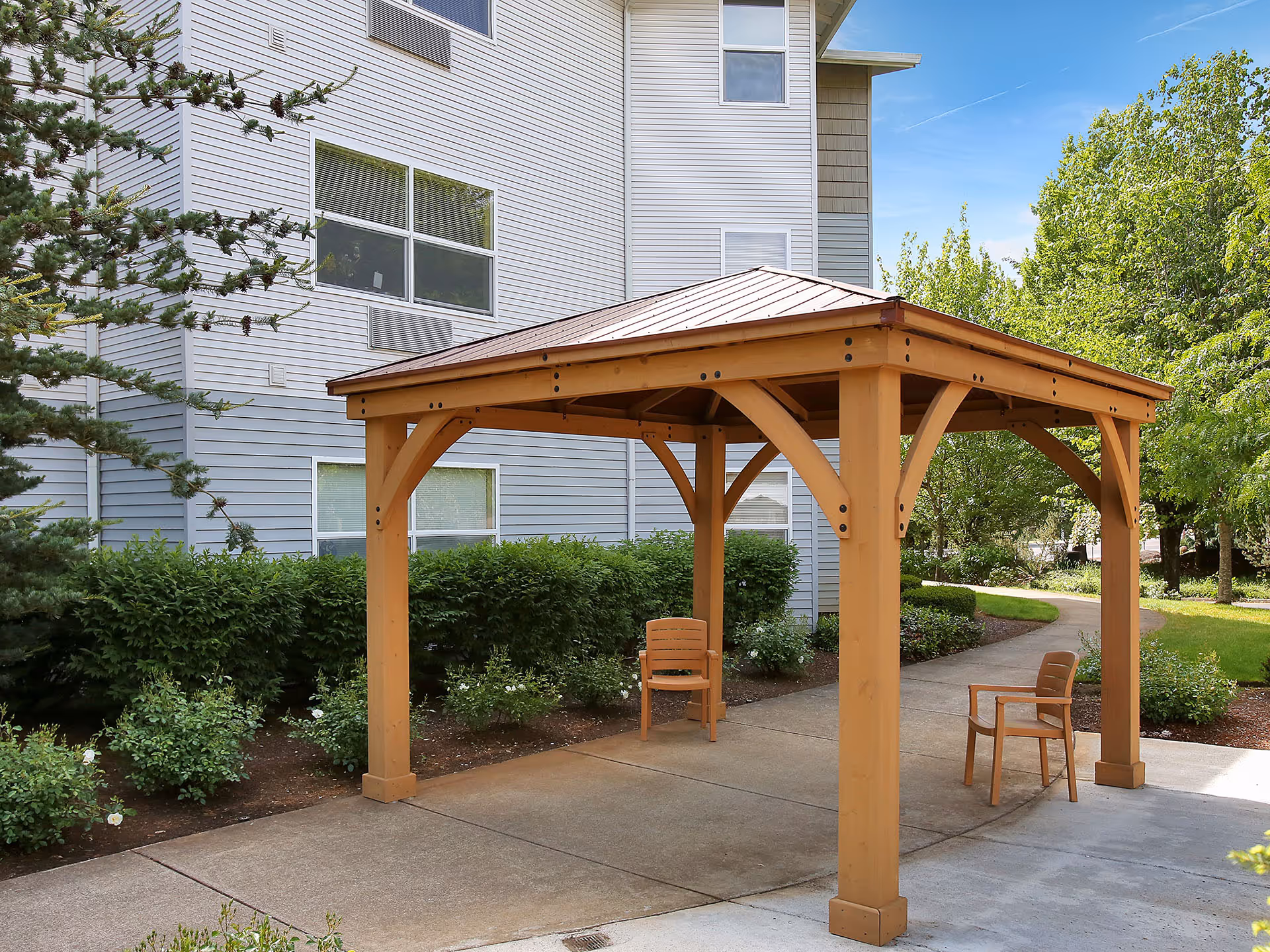 Outdoor wooden gazebo with a metal roof and two wooden chairs underneath, situated on a concrete pathway next to a senior living facility building surrounded by green bushes and trees.