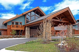 Exterior view of a large building with a peaked roof and stone pillars at the entrance. The building features wooden siding, multiple windows, and a balcony. There is a driveway leading up to the entrance, surrounded by landscaped greenery and small trees under a partly cloudy blue sky.