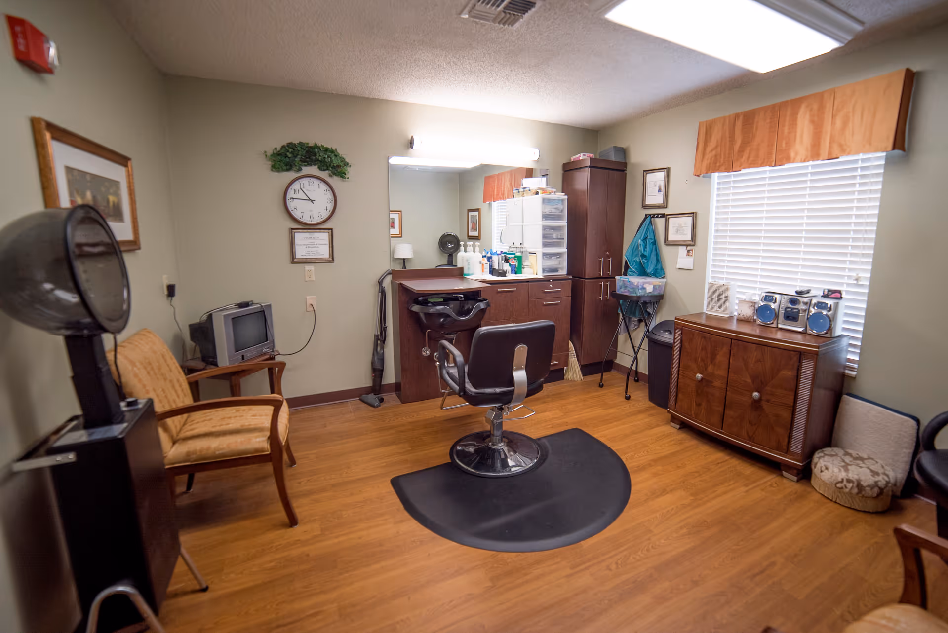 Small hair salon/styling room with a central barber chair, wash sink and mirror, cabinetry, hooded dryer and seating on wood floors.