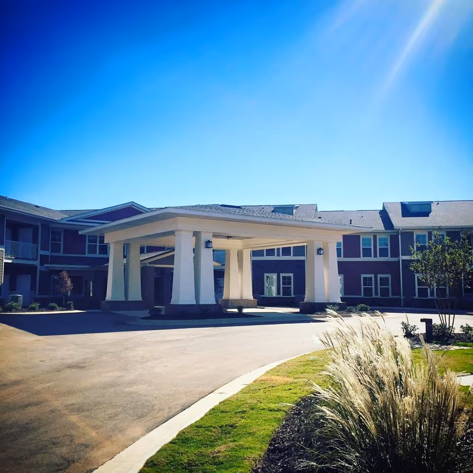 Exterior view of The Lodge At Johns Road showing the entrance driveway with a covered drop-off area supported by large white columns, surrounded by a two-story building with multiple windows under a clear blue sky. There is landscaping with grass and ornamental grasses in the foreground.