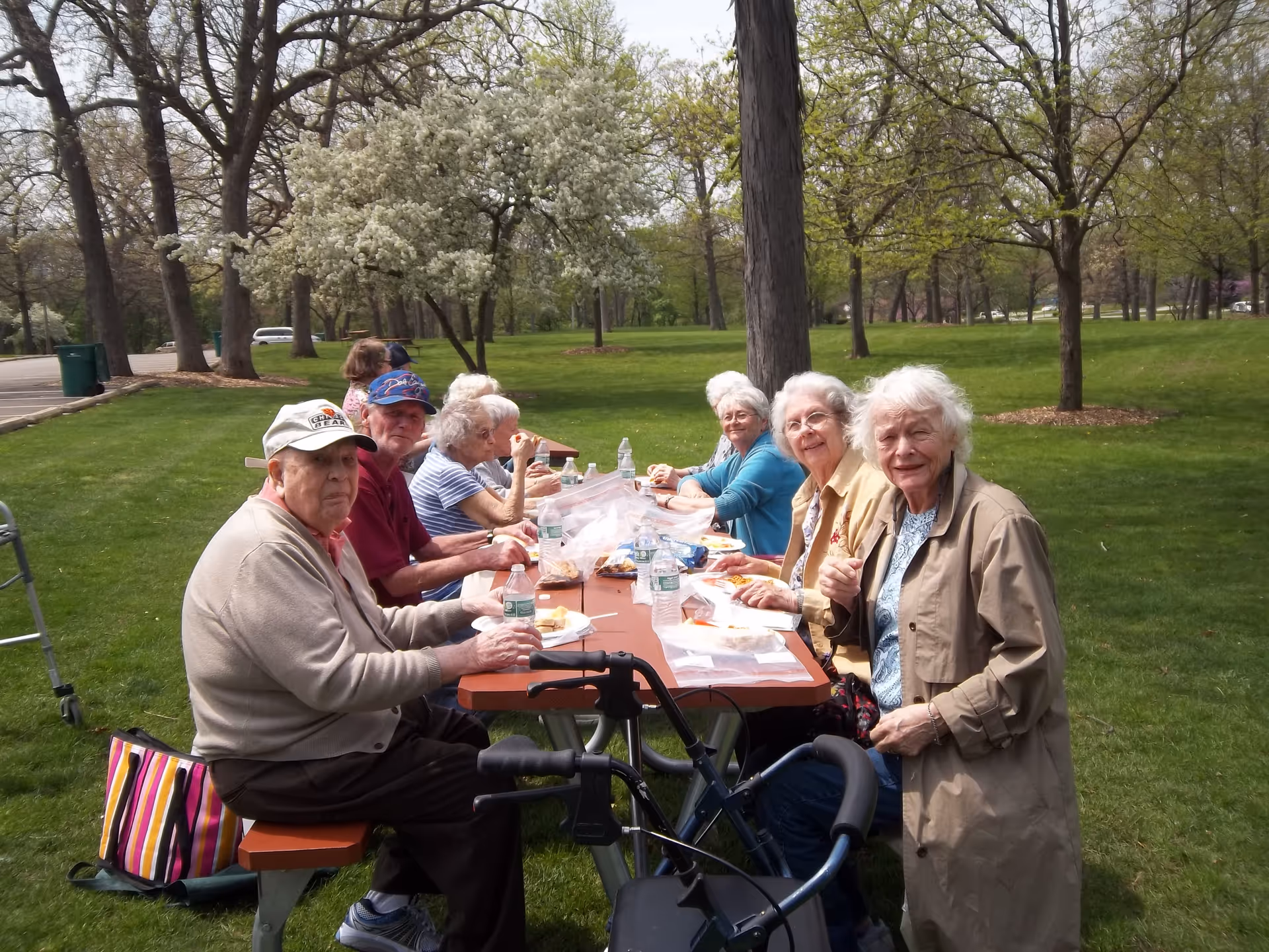 A group of elderly people sitting at a picnic table outdoors in a park-like setting, enjoying a meal together. The table is covered with food items and water bottles. Trees with green leaves and white blossoms surround the area, and a walker is visible in the foreground.