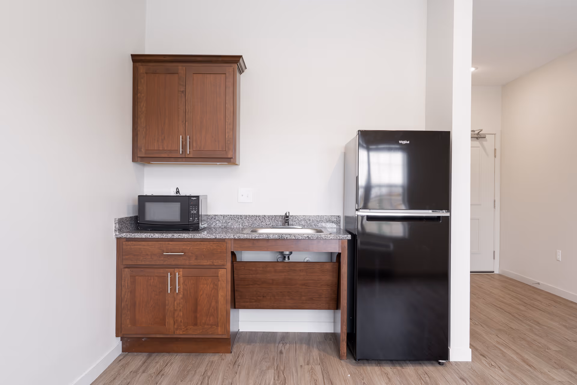 Small kitchenette area with wooden cabinets, a granite countertop, a microwave, a stainless steel sink, and a black Whirlpool refrigerator. The floor is wood laminate and the walls are painted white. A white door is visible in the background.