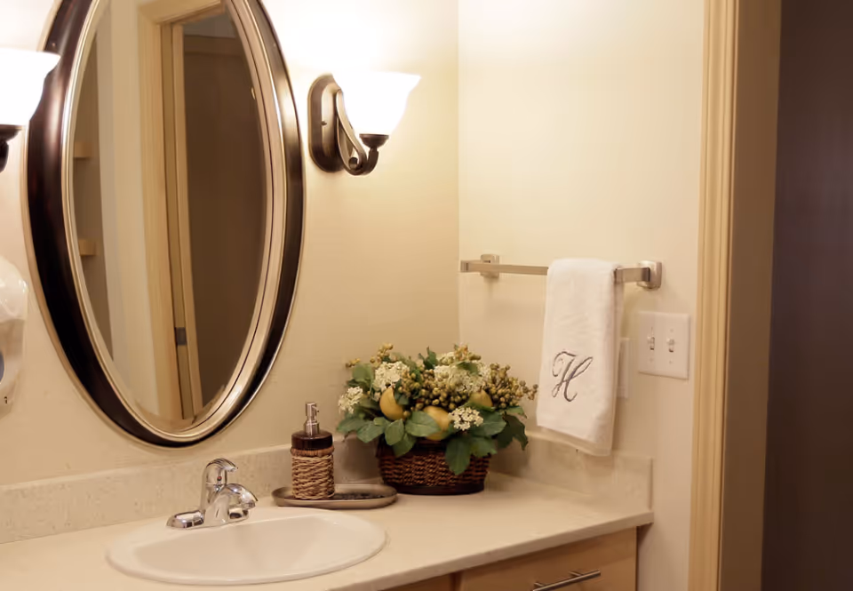 A bathroom vanity area with a white sink, chrome faucet, and beige countertop. Above the sink is an oval mirror with a dark brown frame, flanked by two wall-mounted light fixtures with frosted glass shades. On the countertop, there is a soap dispenser and a decorative basket with green leaves and white flowers. A white hand towel with the letter 'H' embroidered hangs on a towel rack mounted on the wall next to a light switch panel.