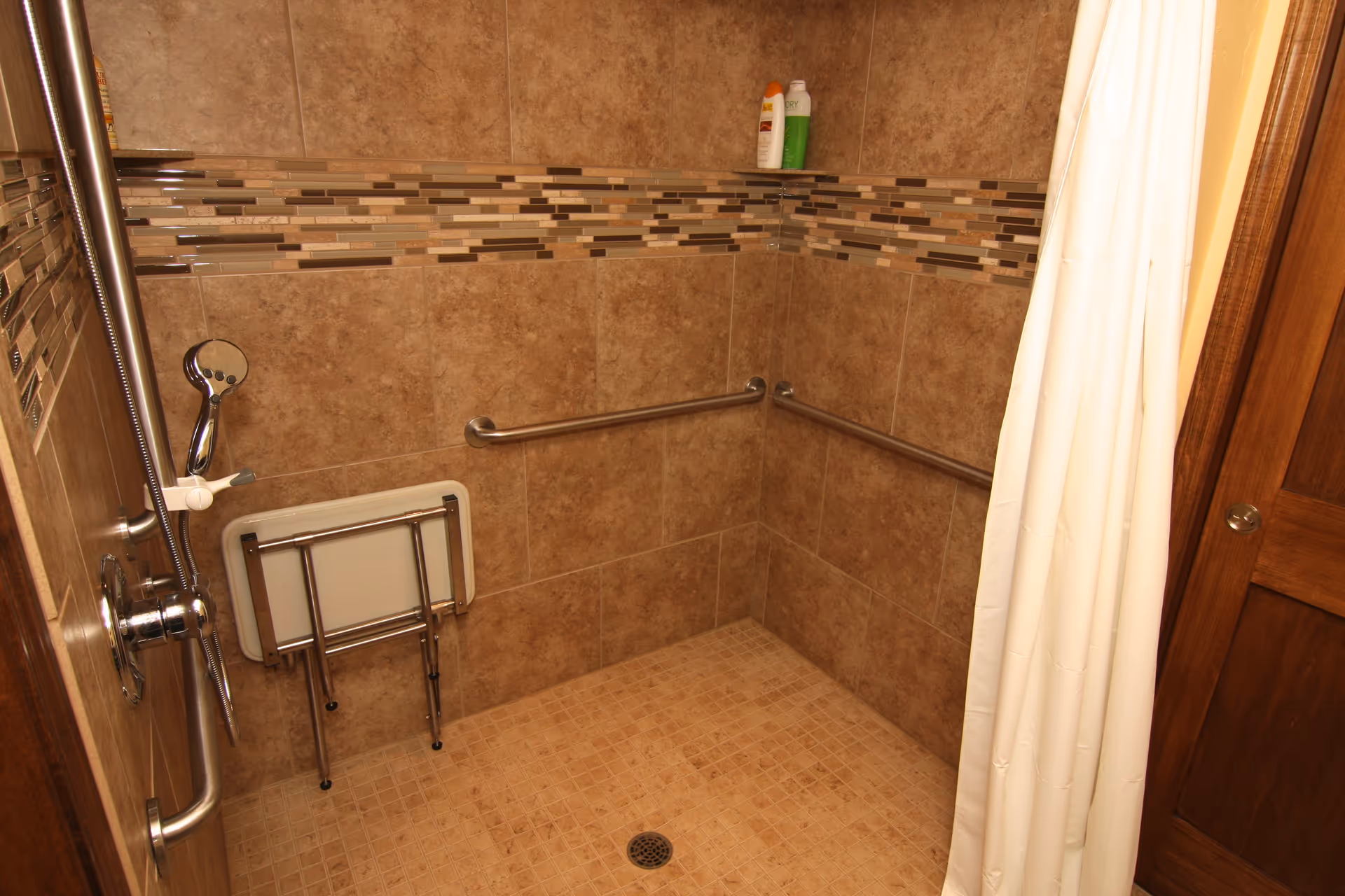 A tiled walk-in shower with beige and brown mosaic accent tiles, equipped with two horizontal grab bars, a handheld showerhead, a foldable shower seat, and a white shower curtain partially drawn to the side. Two bottles of shower products are placed on a corner shelf.