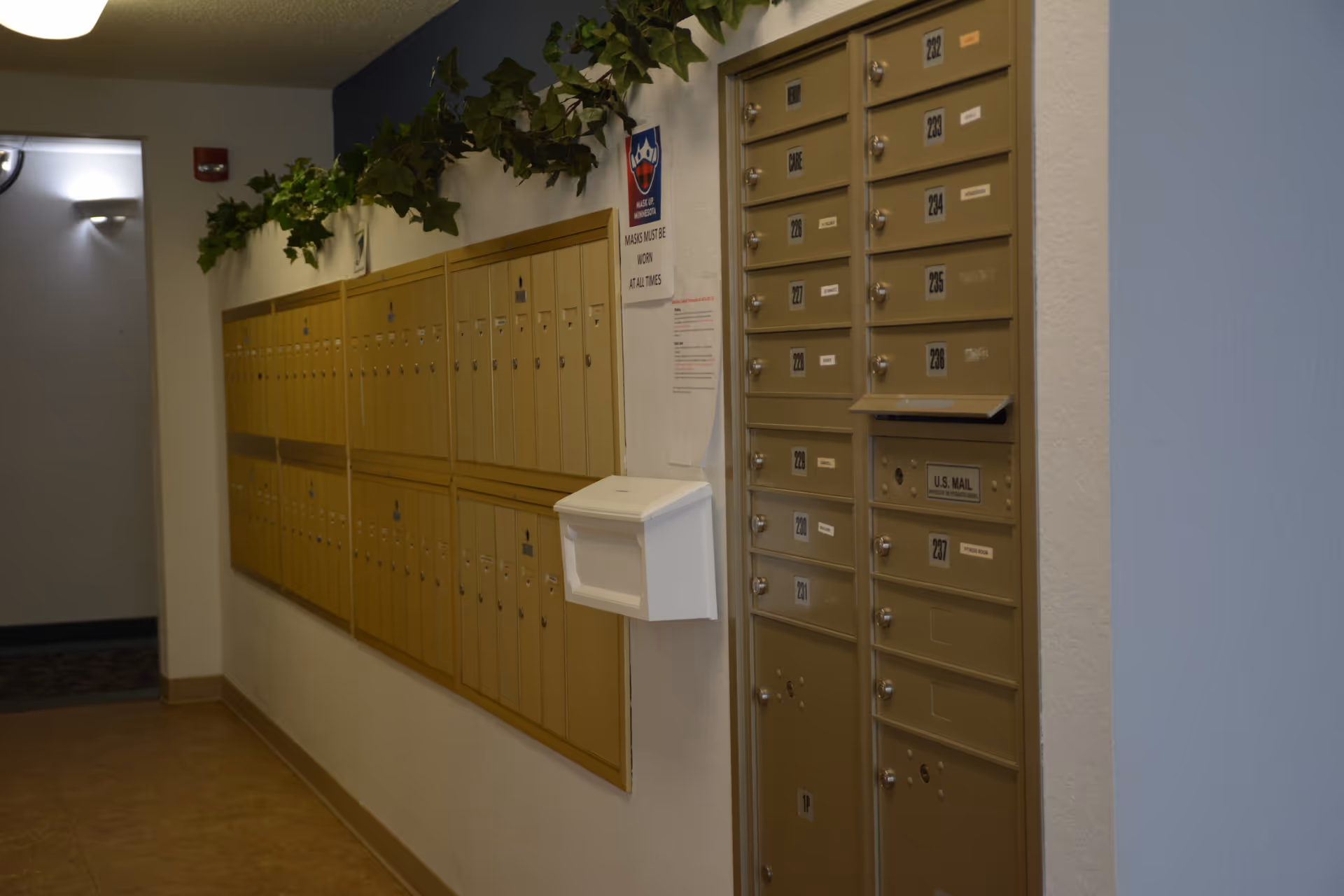 Interior hallway with multiple beige mailboxes mounted on the walls. There is a white mailbox attached to the corner wall and a sign indicating that masks must be worn at all times. Green leafy garlands decorate the top of the mailboxes.