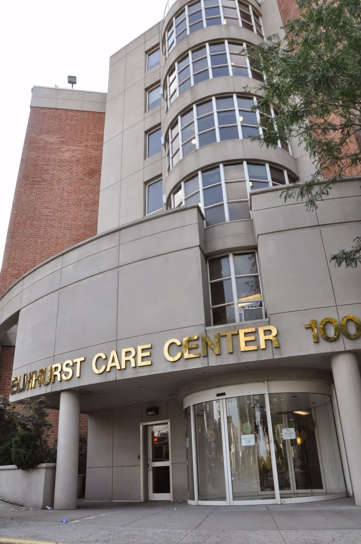 Exterior view of Elmhurst Care Center building entrance with curved facade, large windows, and gold lettering displaying the facility name above the revolving door entrance.