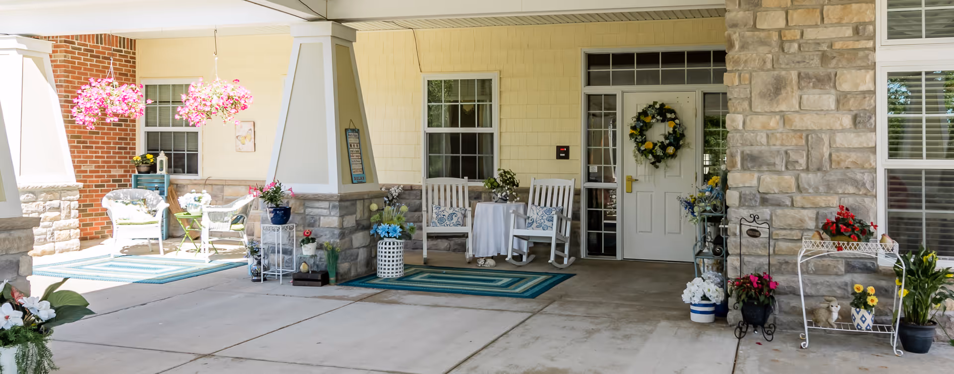 Covered entrance porch with rocking chairs, potted plants, hanging baskets, and a wreath on the door.