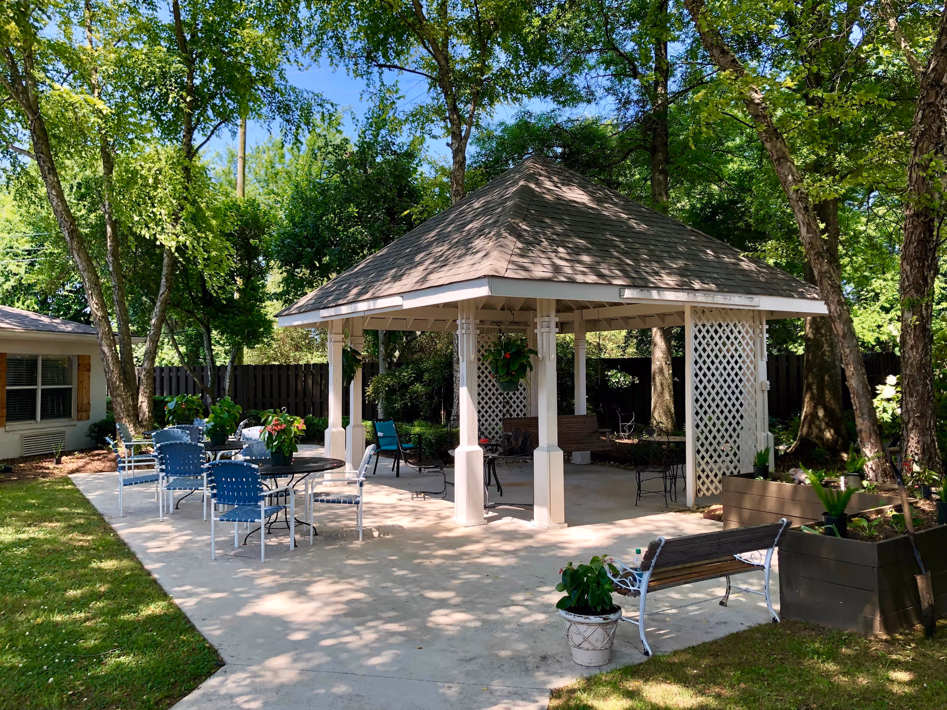 Outdoor gazebo with a shingled roof surrounded by trees and greenery. There are several tables and chairs arranged on a concrete patio, with potted plants on the tables and around the area. A wooden bench and planter boxes are also visible, creating a peaceful garden setting.