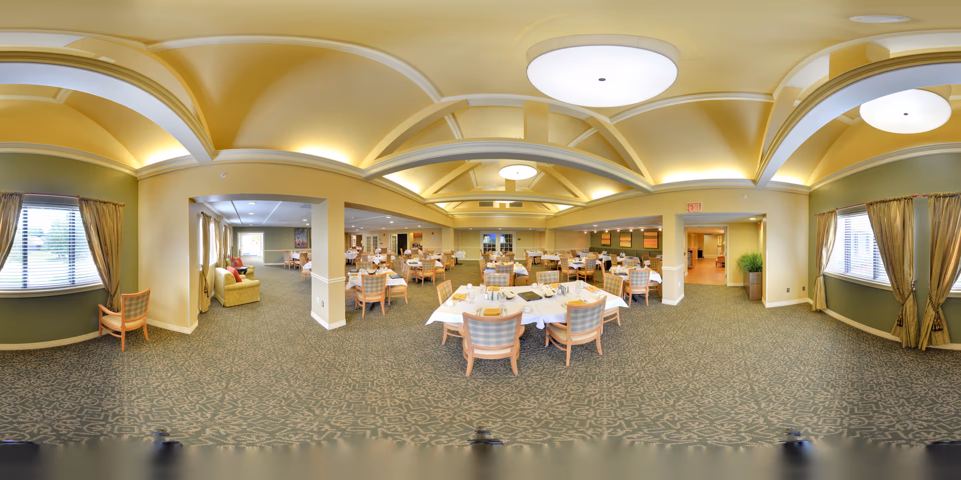 A spacious dining room in a senior living facility with multiple tables covered with white tablecloths and set with plates, glasses, and napkins. The room features patterned carpet, large windows with curtains, soft yellow walls, and a vaulted ceiling with recessed lighting and large circular ceiling lights. There are several chairs around each table and a small seating area with a sofa and chairs near the windows.