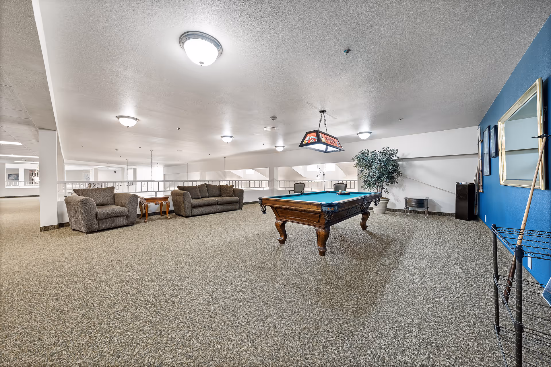 A spacious indoor recreational room with a pool table in the center, a hanging light fixture above it, two gray upholstered sofas with a wooden side table between them, a large potted plant, a blue accent wall with a mirror, and a rack holding pool cues and other accessories.