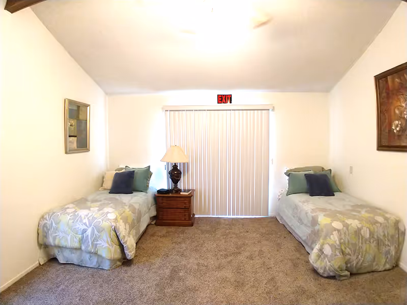 Spacious bedroom with two twin beds separated by a nightstand and lamp facing sliding vertical blinds with an EXIT sign above.
