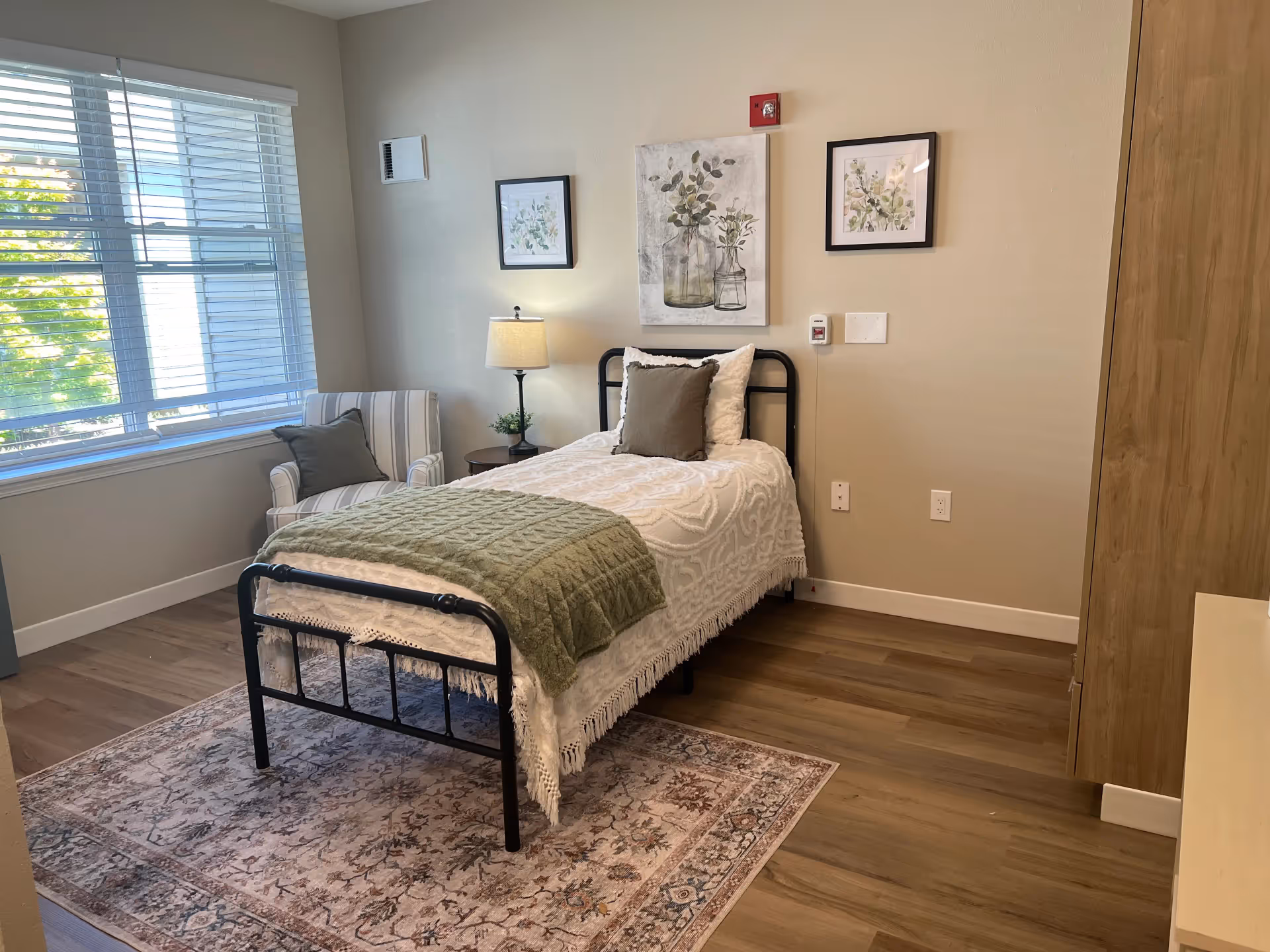 Sunlit furnished single-occupancy bedroom with a bed, armchair, bedside table, and framed wall art.