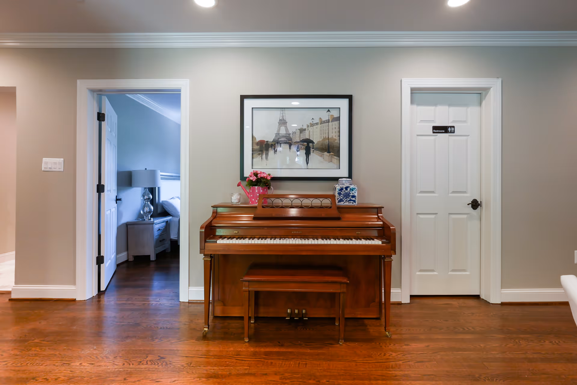 Interior view of a room with a wooden piano and bench against a beige wall. Above the piano is a framed picture of people walking near the Eiffel Tower. To the left is an open doorway leading to a bedroom with a bedside table and lamp. To the right is a closed door labeled 'Restrooms'. The floor is wooden.
