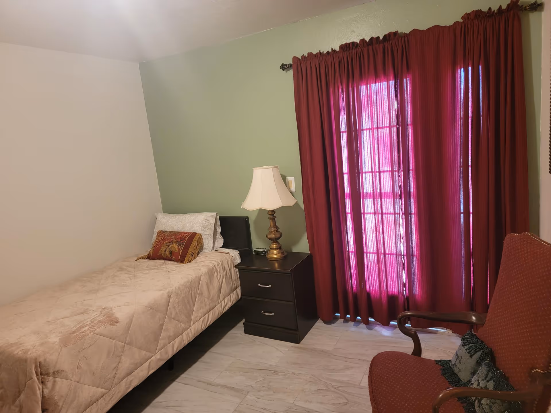 A small bedroom with a single bed covered in a beige quilt and a decorative pillow. Next to the bed is a dark wooden nightstand with two drawers, holding a brass table lamp and a small clock. The room has light-colored walls with one wall painted green. There is a window or door covered by red curtains, and a red upholstered armchair with a decorative pillow is positioned near the window.