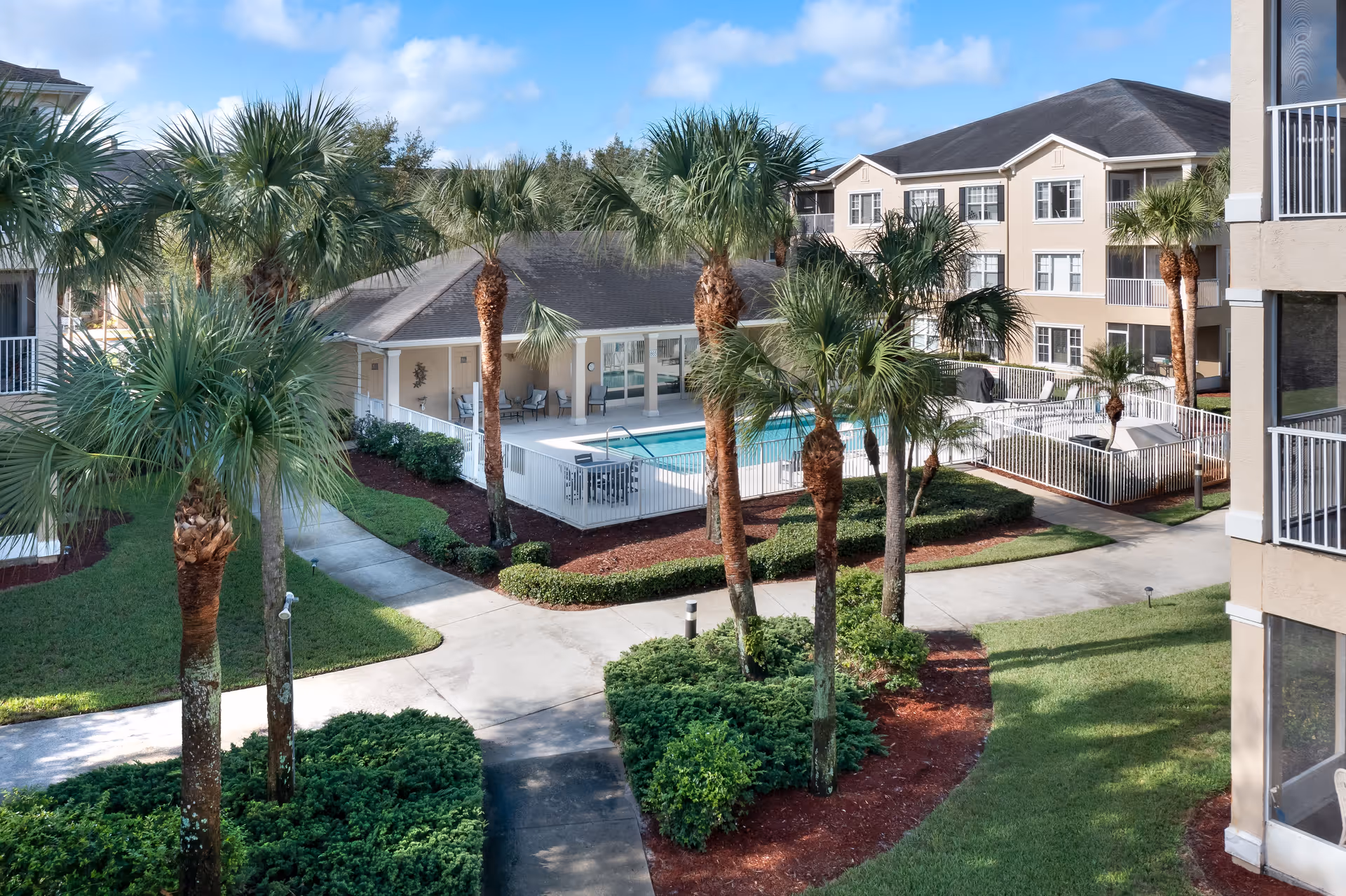 Outdoor view of a senior living facility courtyard with palm trees, manicured bushes, and a swimming pool enclosed by a white fence. Surrounding the courtyard are multi-story beige buildings with balconies and windows under a blue sky with some clouds.