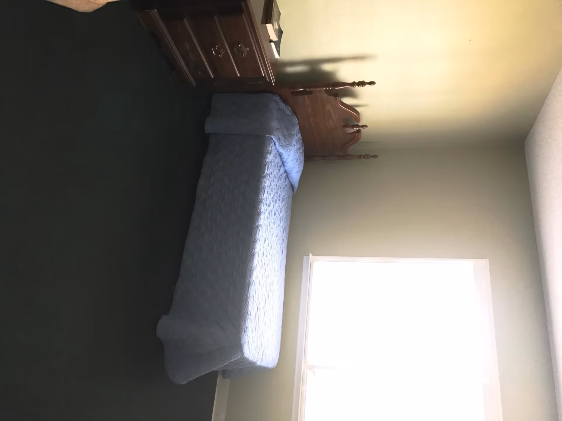 A simple bedroom with a single bed covered in a light blue quilted bedspread. Next to the bed is a wooden nightstand with two drawers and a few items on top. The room has light-colored walls and a window letting in bright natural light.