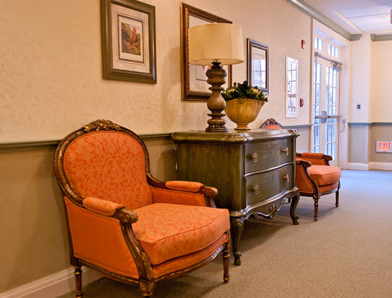A hallway in a senior living facility with two ornate orange upholstered armchairs on either side of a dark wooden chest of drawers. On top of the chest is a decorative lamp and a plant. The walls are decorated with framed artwork, and there is a glass door at the end of the hallway letting in natural light.